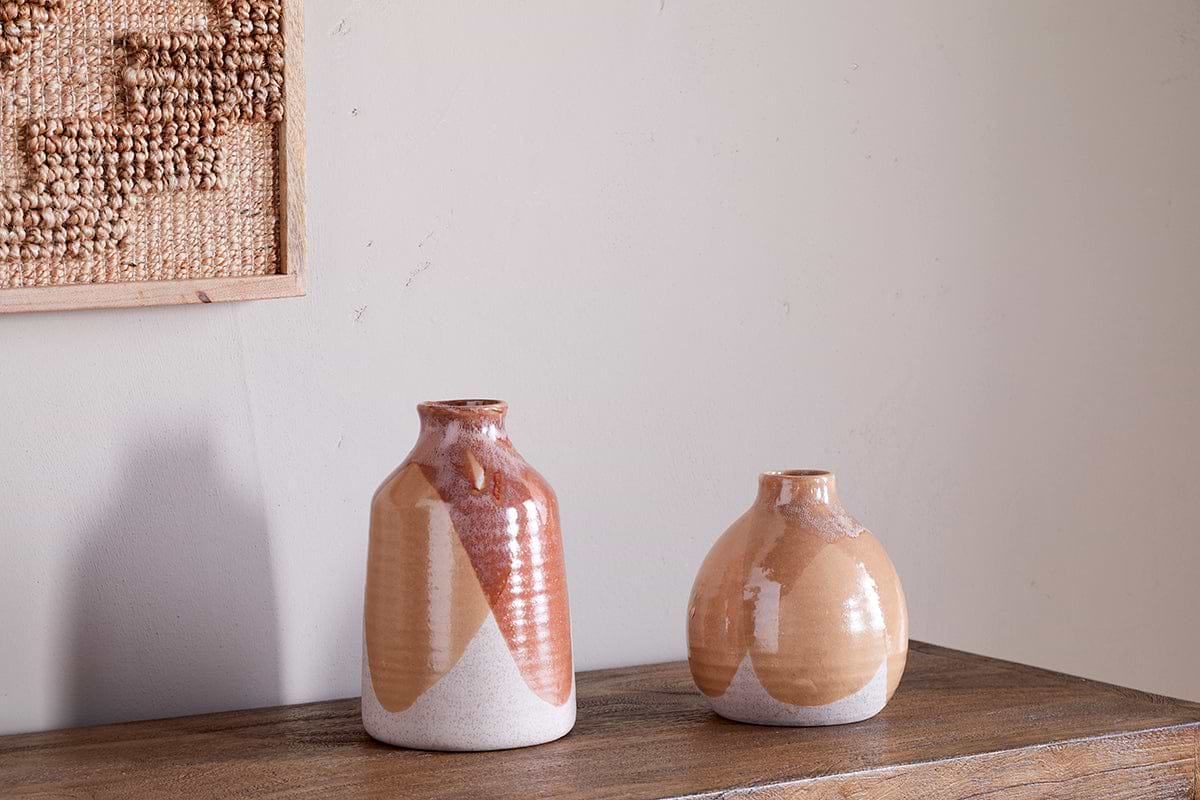 Two nkuku Lucia Ceramic Vases in a terracotta mix with geometric patterns rest on a wooden surface against a light wall. A framed woven textile to the left adds texture to the minimalist decor.