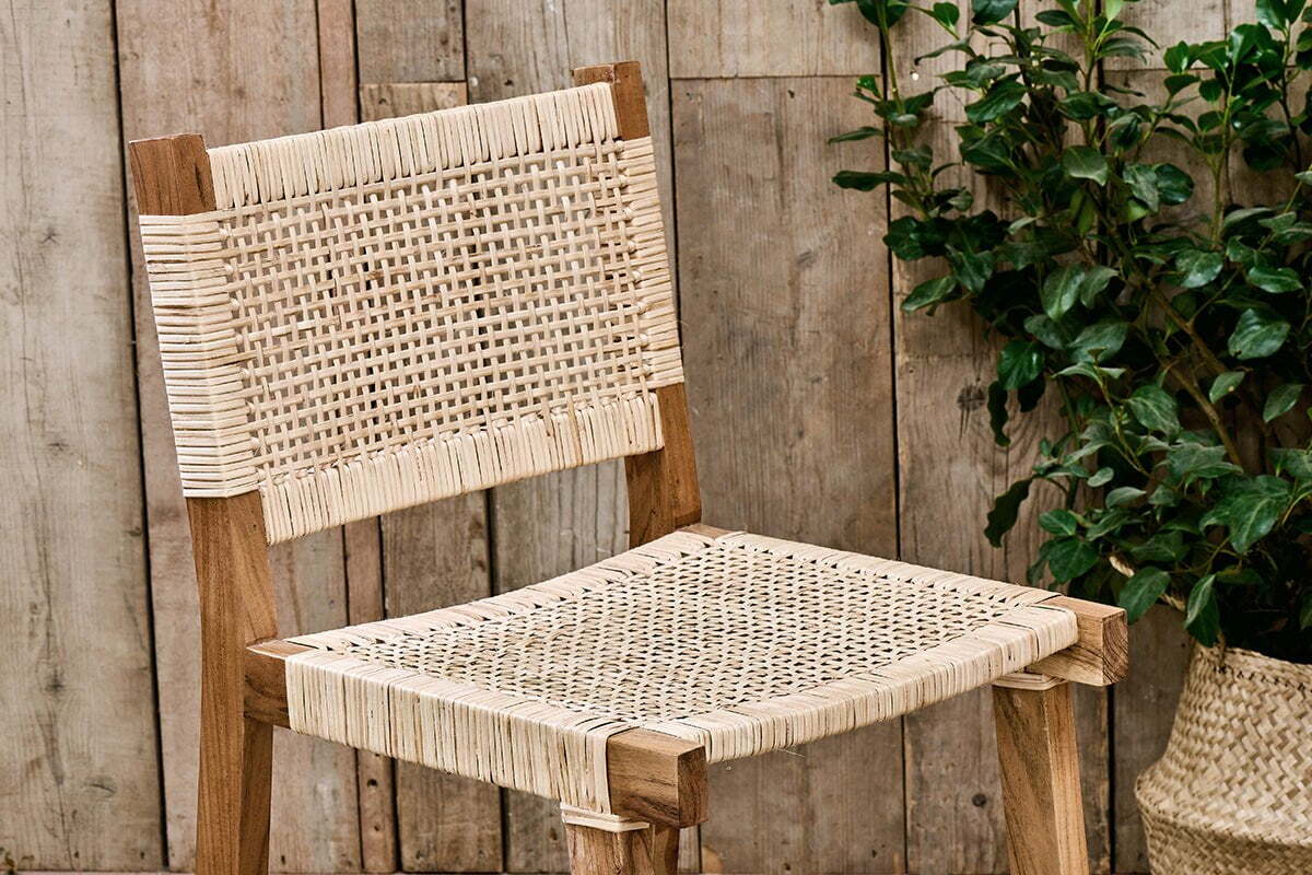 The Madrisana Acacia & Rattan Bar Stool by nkuku, featuring a woven light-colored seat and backrest, is placed before rustic wooden paneling. A potted green plant appears partially on the right side of the image.