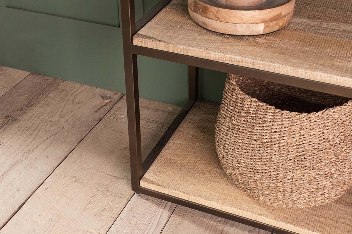 A close-up of nkuku's Marjori Mango Wood And Iron Standing Shelves - Wide displays a black metal frame with mango wood shelves. A round wooden bowl sits on the middle shelf and a large woven basket is on the shelf below, atop wide, weathered wooden planks.