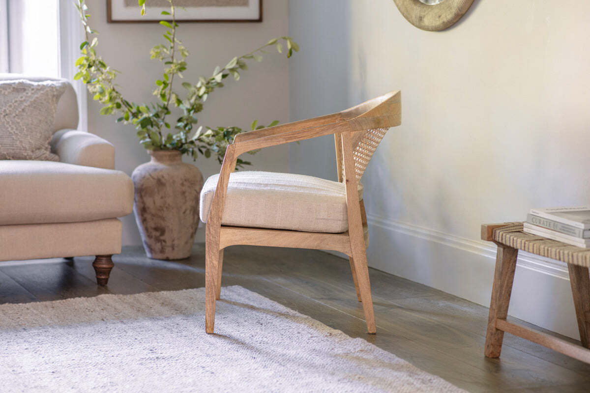 The nkuku Nabhi Mango Wood & Cane Occasional Chair features an upholstered cotton cushion and sits on a light rug. A beige sofa with a textured pillow is to the left, and a large textured vase with greenery stands behind the sofa.