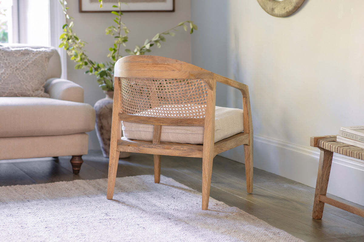 The Nabhi Mango Wood & Cane Occasional Chair by nkuku, featuring a natural mango wood frame and cane back with an upholstered cotton cushion, is shown from behind in a bright living room with a beige sofa, green plant, rug, and side table.
