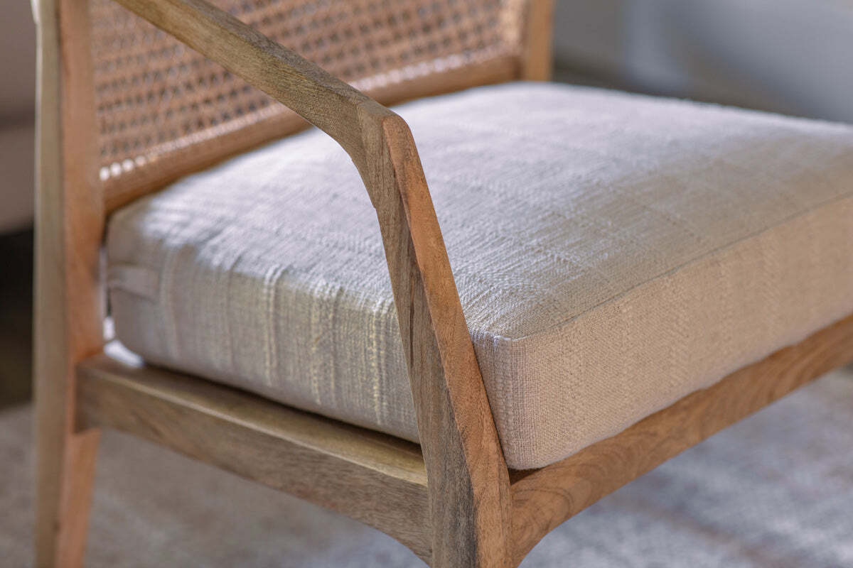 Close-up of the Nabhi Mango Wood & Cane Occasional Chair by nkuku, featuring a natural wood frame, handcrafted cane back, and thick cream linen cushion. Curved armrest showcases its mid-century modern elegance.