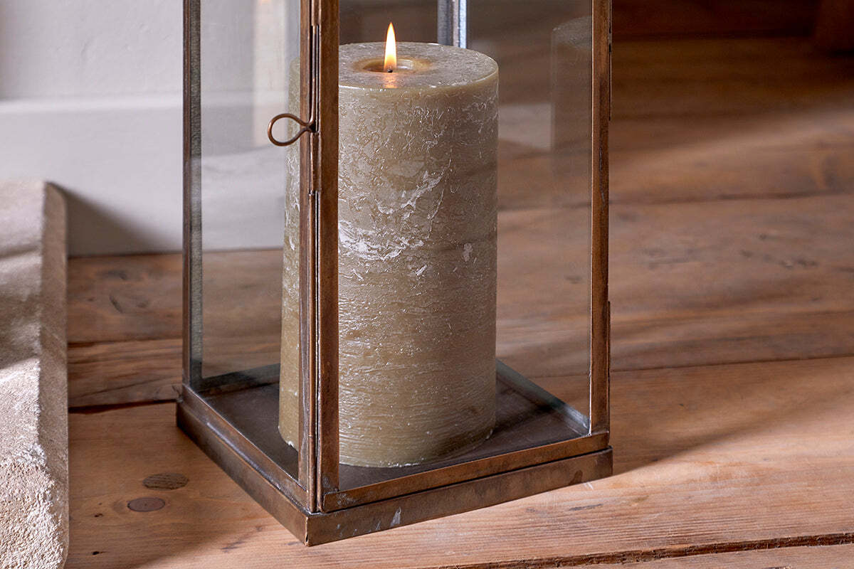 The Nivas Lantern - Antique Brass by nkuku sits on a wooden floor, glowing with a steady pillar candle. Warm natural light highlights this elegant Eastern-style home accessory.
