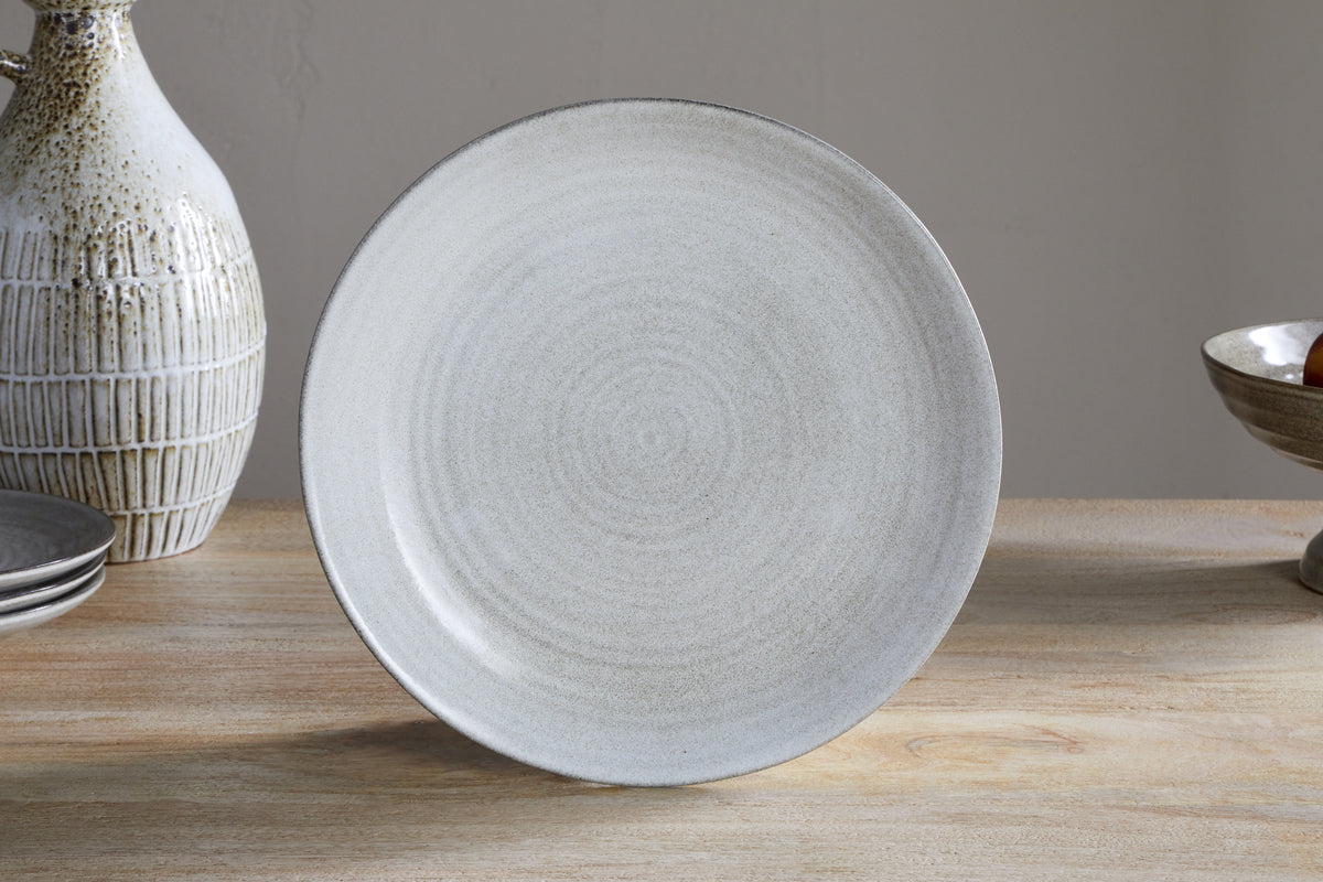 The Valinha Earthenware Dinnerware Set - Grey by nkuku, featuring a round, light gray ceramic plate with a subtle textured pattern and reactive glaze, is shown upright on a wooden table among other serveware and pottery.