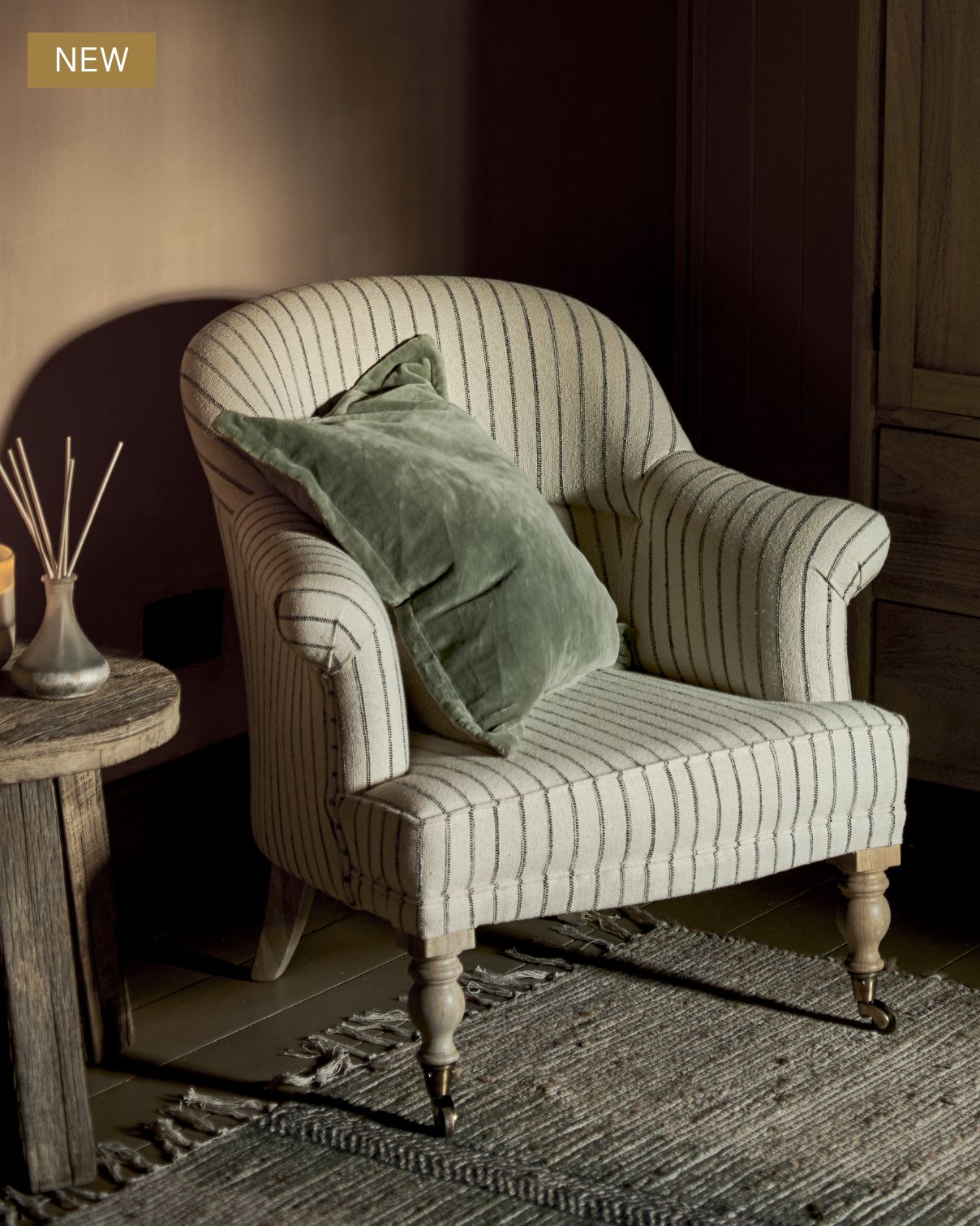 A cream, striped upholstered armchair with wooden legs holds a green velvet pillow. It sits on a textured rug next to a small round table with reed diffusers in a softly lit, cozy room.