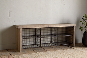 The nkuku Abha Reclaimed Wood & Iron Storage Bench – Natural is a versatile entryway organizer featuring six black wire baskets for shoe storage, set on a wooden floor beside a potted green plant.