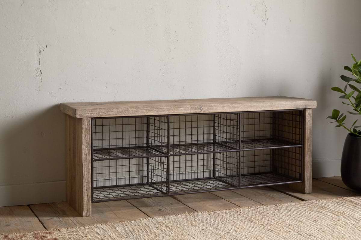 The nkuku Abha Reclaimed Wood & Iron Storage Bench – Natural is a versatile entryway organizer featuring six black wire baskets for shoe storage, set on a wooden floor beside a potted green plant.