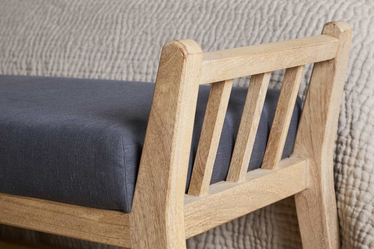 Close-up of the nkuku Avanthi Upholstered Sleigh Bench in charcoal, featuring a cushioned seat and slatted wooden backrest, set before a textured beige quilted blanket.