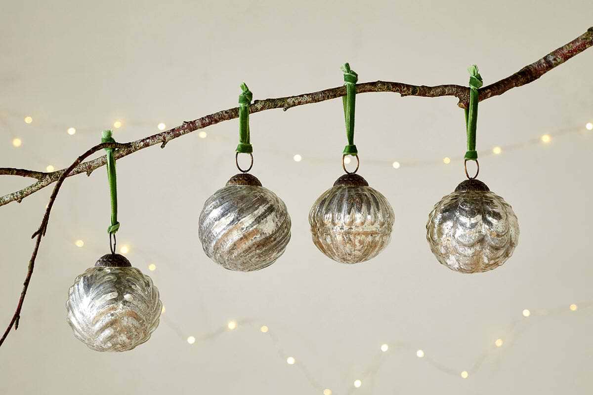 The Adoya Baubles - Aged Nickel (Set of 4) by nkuku hang from a branch with a green velvet tie, surrounded by warm white fairy lights and a soft backdrop. Their ribbed texture adds cozy charm to your festive décor.