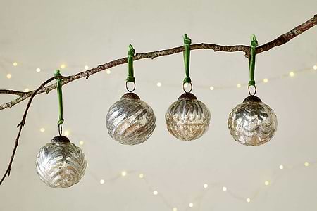 The Adoya Baubles - Aged Nickel (Set of 4) by nkuku hang from a branch with a green velvet tie, surrounded by warm white fairy lights and a soft backdrop. Their ribbed texture adds cozy charm to your festive décor.