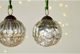 The Adoya Baubles - Aged Nickel (Set of 4) by nkuku hang from green velvet ties, with textured patterns. Warm fairy lights glow softly in the blurred background, adding a cozy and festive touch to your décor.