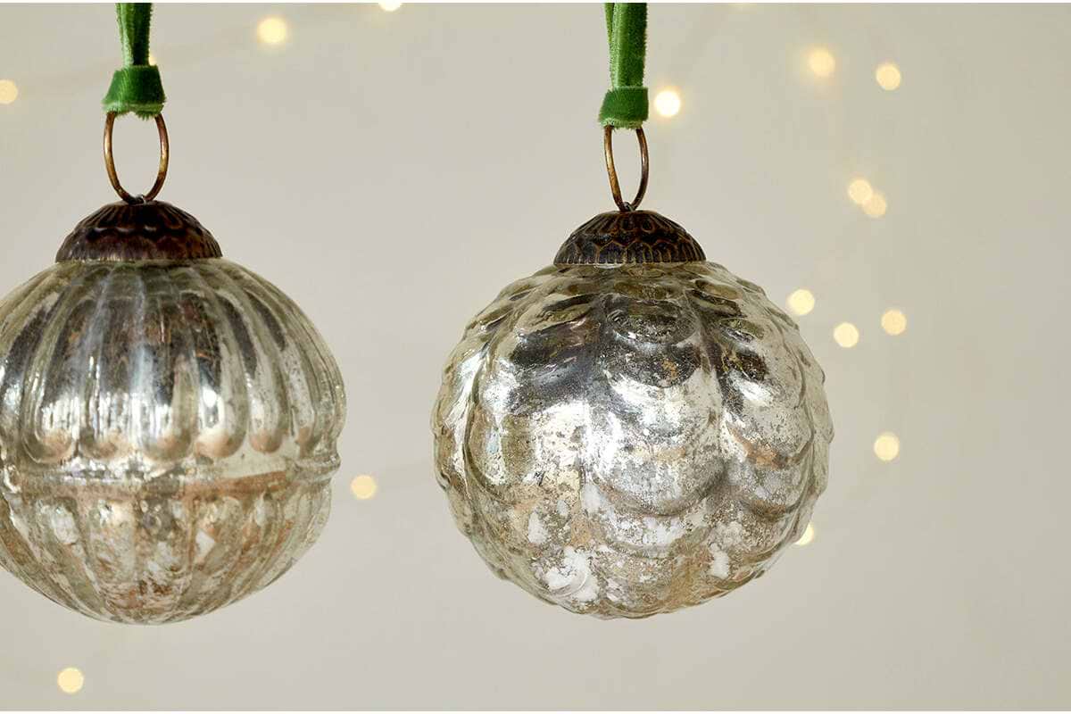The Adoya Baubles - Aged Nickel (Set of 4) by nkuku hang from green velvet ties, with textured patterns. Warm fairy lights glow softly in the blurred background, adding a cozy and festive touch to your décor.