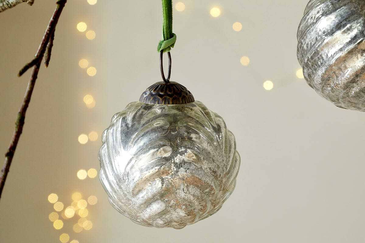 The Adoya Baubles - Aged Nickel (Set of 4) by nkuku, featuring vintage-style ribbed glass with a leafy pattern and a green velvet tie, hang amid festive decor with soft yellow fairy lights for a cozy holiday ambiance.