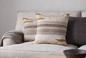 A beige sofa features two nkuku Aravali Wool Cushion Covers in ochre and earthy tones, a gray textured blanket over the arm, a dark gray pillow behind, and a wooden bowl on a light-colored table in front.