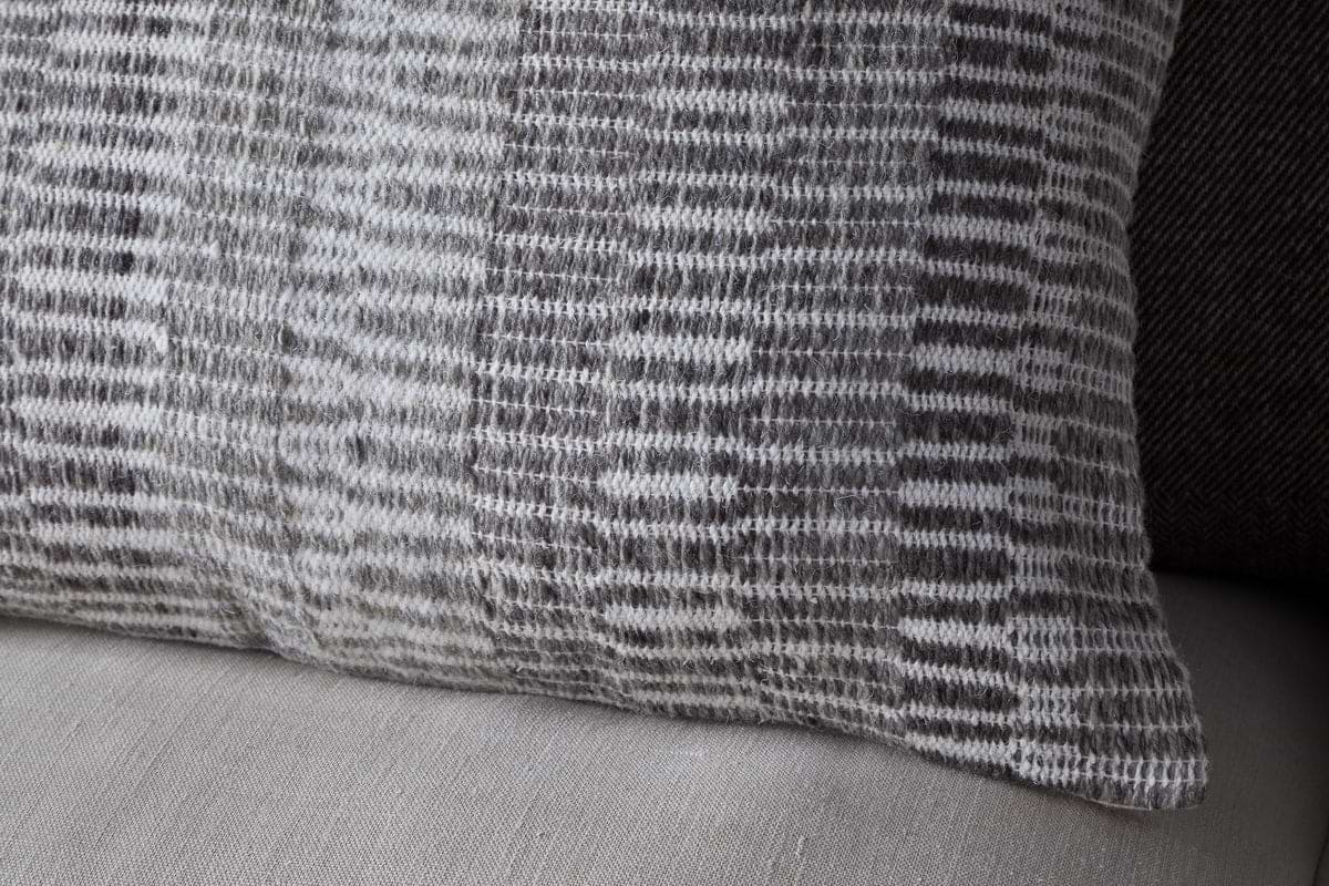 A close-up of the nkuku Aahna Wool Cushion Cover in charcoal, featuring a textured woven design in gray and white wool and recycled polyester, displayed on a light beige fabric surface.