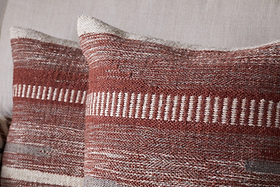 Two Aakash Wool Cushion Covers in earthy rust tones by nkuku, featuring textured horizontal and vertical woven stripes, add warmth and charm as decorative pillows on a light-colored sofa.