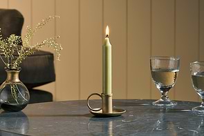 An Amri Iron Candlestick by nkuku in antique brass holds a lit green dinner candle on a round marble table, accompanied by two glasses of water and a small vase of dried flowers. In the background are a dark sofa and light vertical paneling.