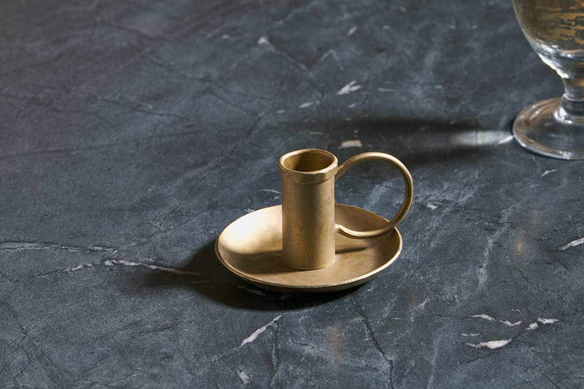 The Amri Iron Candlestick by nkuku, finished in antique brass, sits on a matching round dish atop dark veined marble. To the right, the base of a partially visible goblet or glass casts a soft shadow.