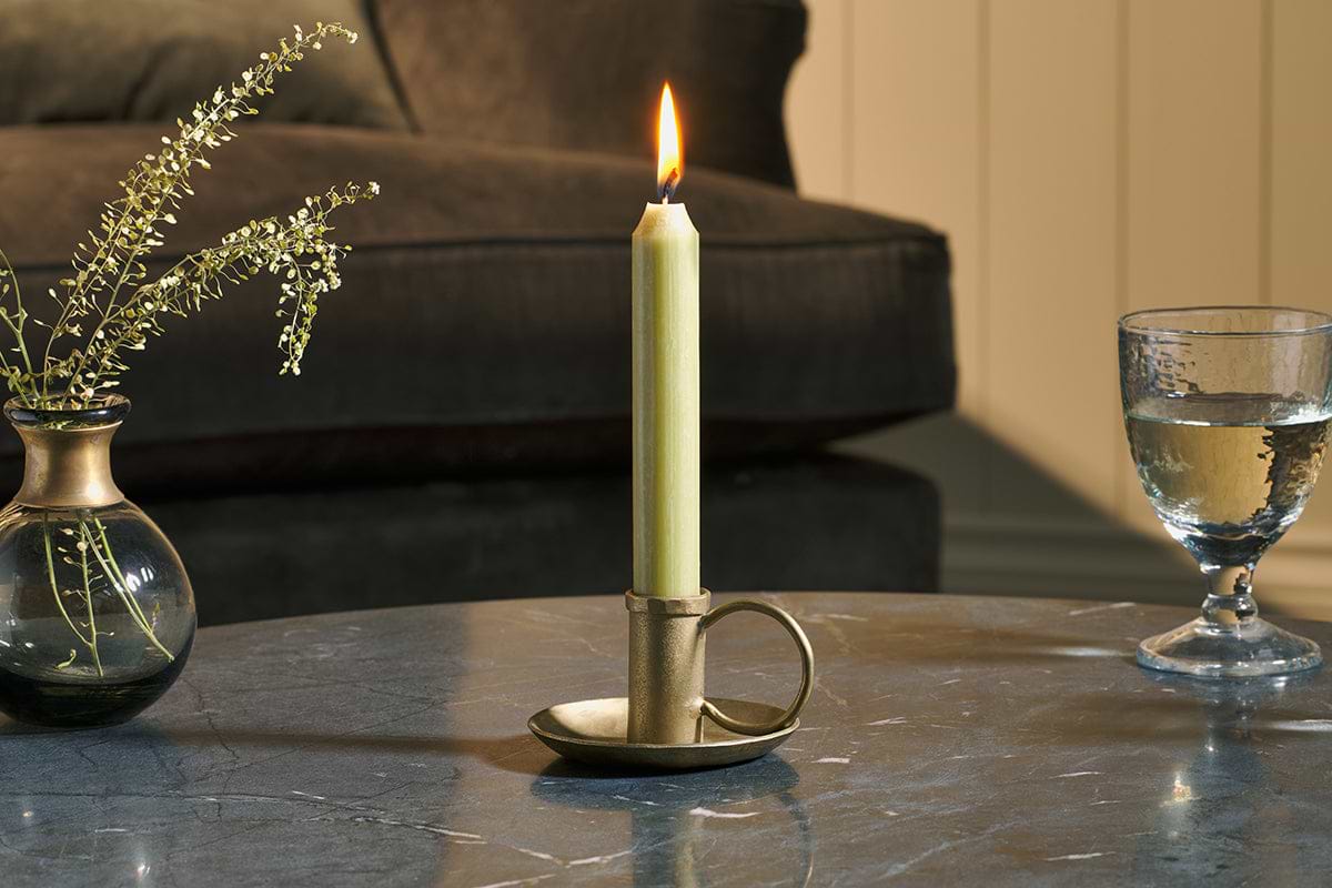 An Amri Iron Candlestick in Antique Brass by nkuku sits lit on a gray marble table, beside a small glass vase with sprigs and a half-filled water glass. A dark sofa and beige paneled wall create a cozy, warm atmosphere.
