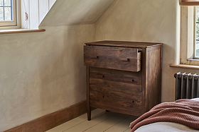 The nkuku Anbu Acacia Chest of Drawers in Washed Walnut stands in a cozy room with cream walls, wood trim, a radiator, and part of a bed with a rust-colored textured blanket visible. The top drawer is partially open.