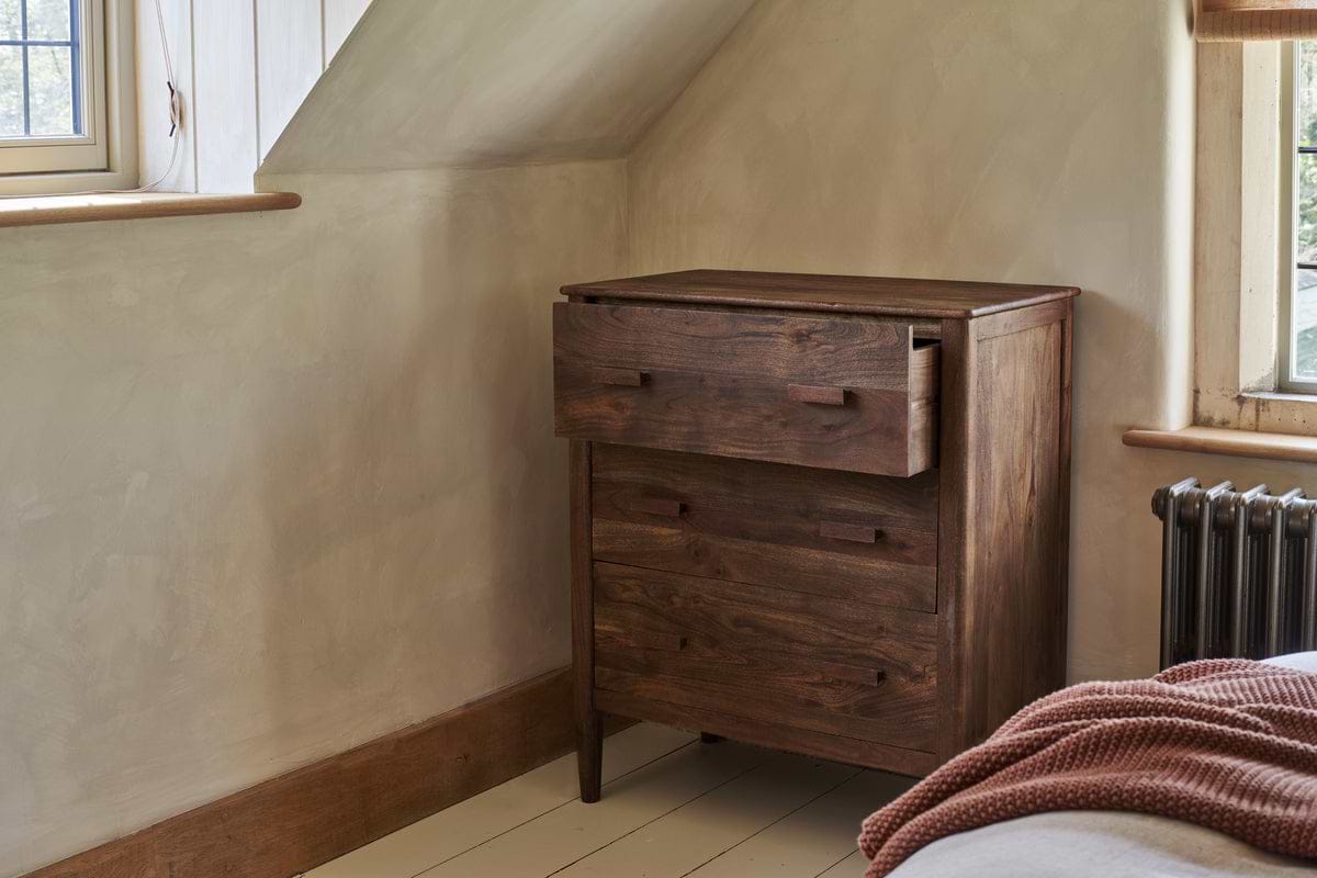 The nkuku Anbu Acacia Chest of Drawers in Washed Walnut stands in a cozy room with cream walls, wood trim, a radiator, and part of a bed with a rust-colored textured blanket visible. The top drawer is partially open.