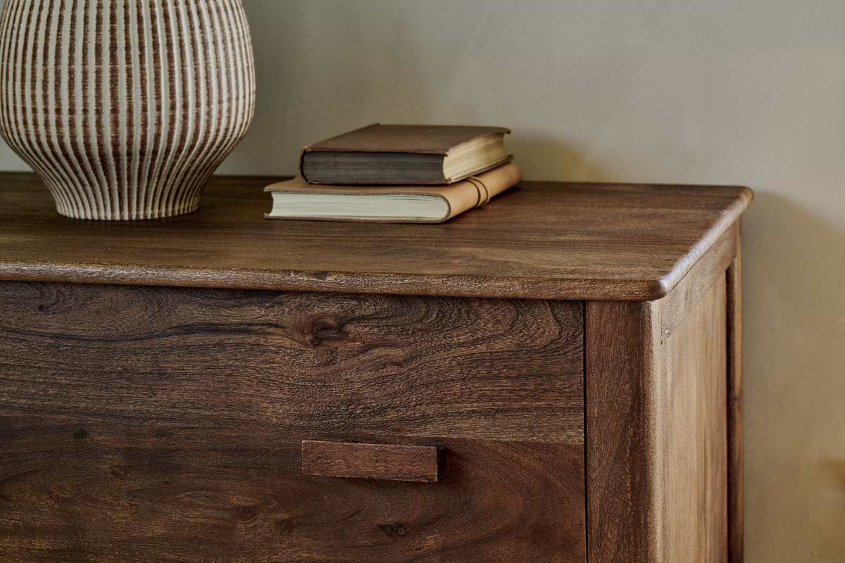 A close-up of the Anbu Acacia Chest of Drawers – Washed Walnut by nkuku features a striped ceramic vase and three stacked books on top, set against a neutral wall background.