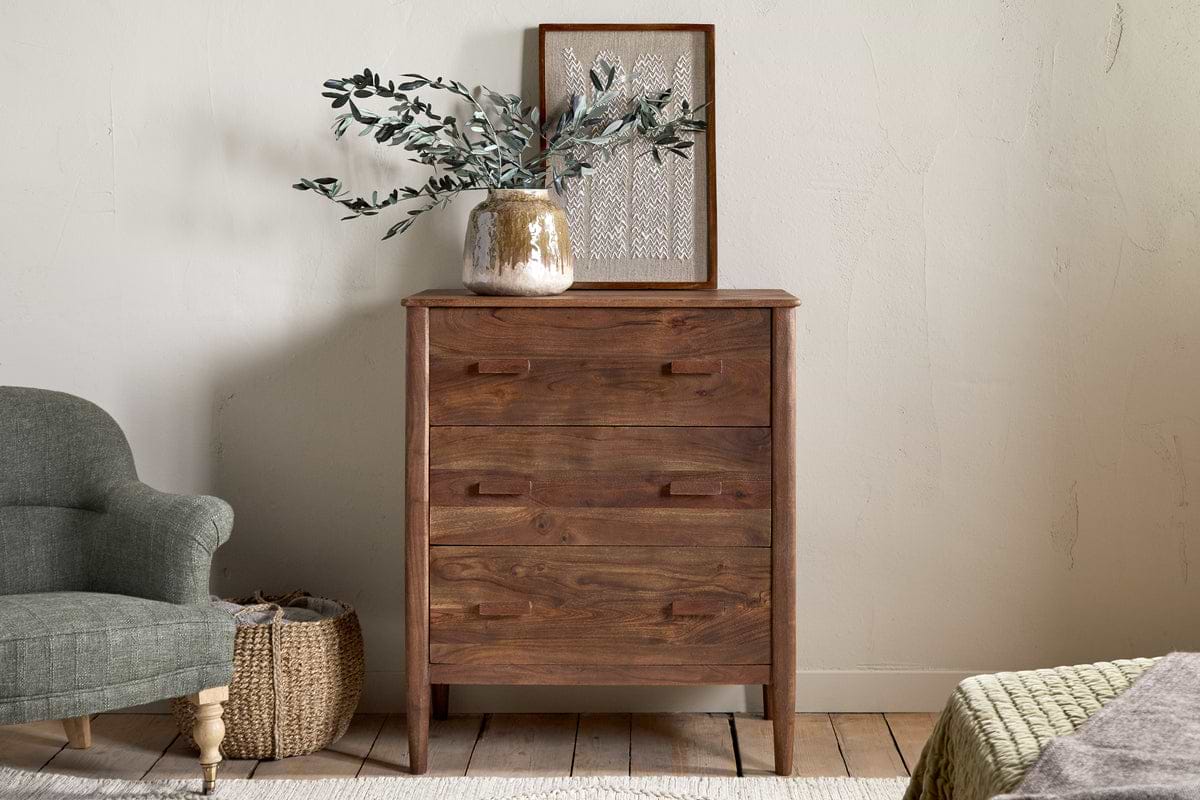 The Anbu Acacia Chest of Drawers by nkuku in washed walnut stands against a light wall, topped with a decorative vase and framed artwork, while a gray armchair and woven basket add style and storage to the bedroom.