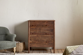 The nkuku Anbu Acacia Chest of Drawers - Washed Walnut, featuring three handles, stands on a rug against a light textured wall. Nearby are a green armchair, woven basket, and part of a bed in this cozy bedroom storage area.
