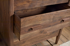 The nkuku Anbu Acacia Chest of Drawers in Washed Walnut features a close-up of its natural wood grain, rectangular handles, and wooden legs next to a textured rug—ideal bedroom storage with an open drawer showcasing its elegant design.