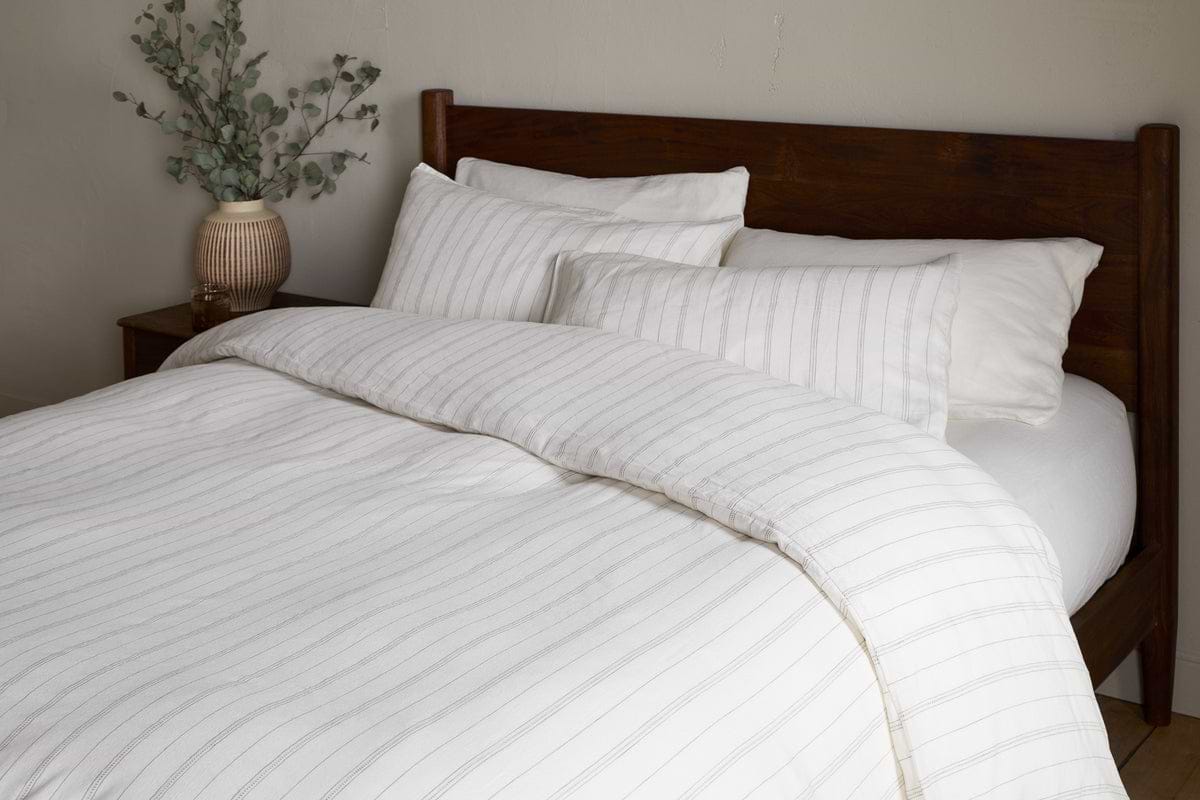 A neatly made bed with white and light gray striped bedding, four pillows, and a wooden headboard features the nkuku Anta Hemp & Cotton Duvet Cover in Sage Green. A ceramic vase with green branches sits on the bedside table.