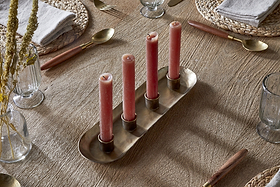 A wooden dining table set for a meal features woven placemats, gold utensils with wooden handles, clear glasses, gray napkins, and the nkuku Amita Long Candle Holder in antique brass as a centrepiece holding four pink candles. Vases with dried grasses complete the scene.