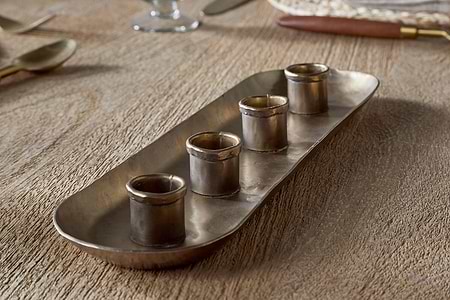 The nkuku Amita Long Candle Holder in antique brass features a rectangular design with rounded corners and four small cylindrical cups in a row, creating an elegant centrepiece for your dining table.