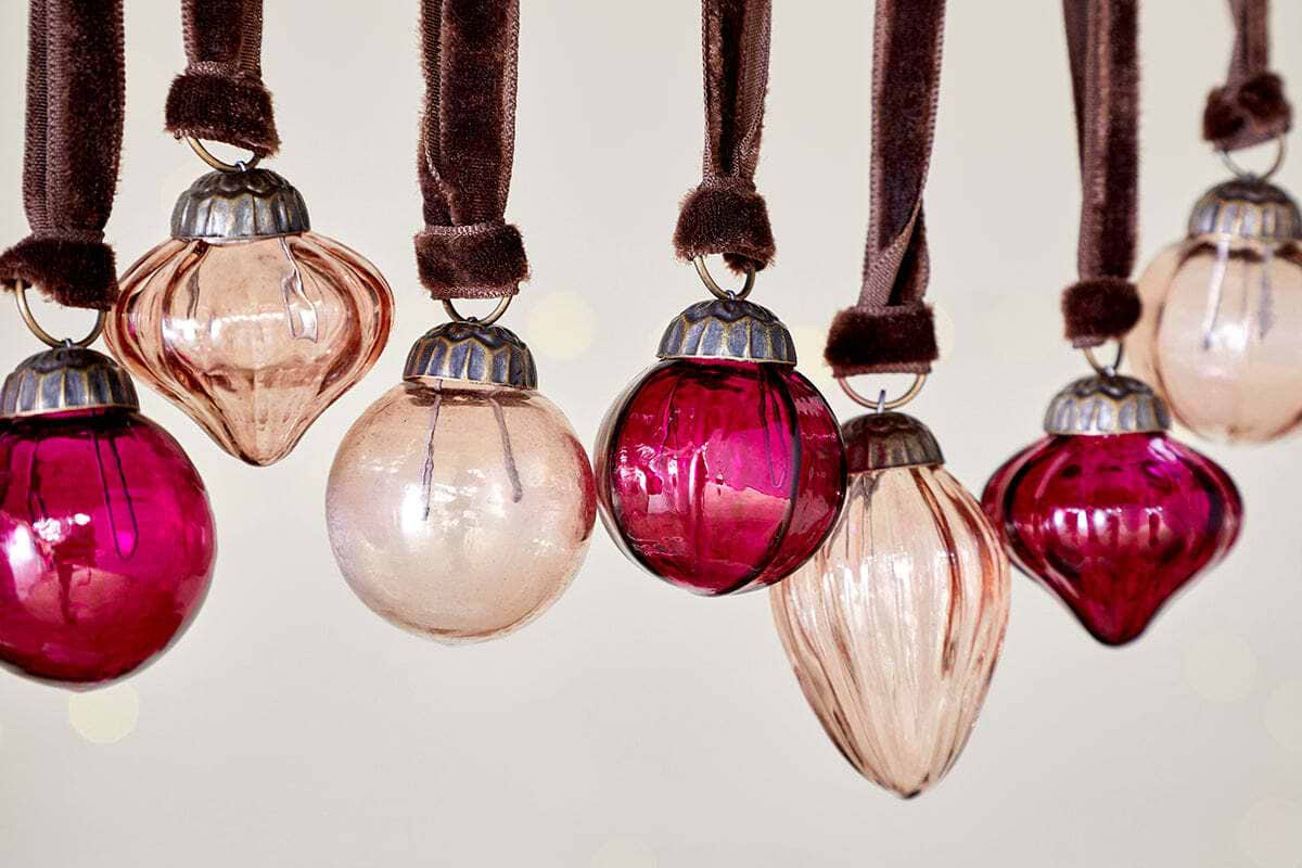 A row of nkuku Adisa Bauble Jar - Ruby & Copper ornaments (set of 16) in pink and red hues hangs from brown velvet ribbons, featuring assorted vintage-style shapes with silver metal caps.