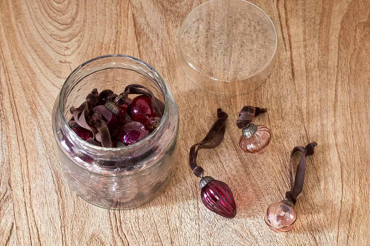 The nkuku Adisa Bauble Jar - Ruby & Copper (Set of 16) features mini glass baubles in festive shades, stored in a recycled glass jar. Some ornaments sit outside the open jar, making it perfect for adding holiday charm to any space.