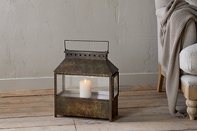 The Akoko Wide Lantern by nkuku, featuring a brown patina and glowing white pillar candle, adds rustic charm beside a beige upholstered chair with a knitted blanket against a textured light-toned wall.