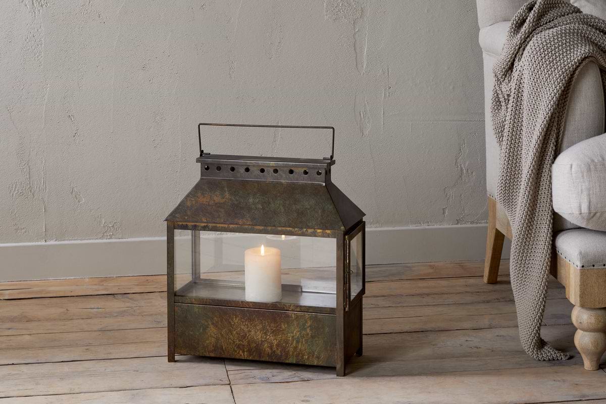 The Akoko Wide Lantern by nkuku, featuring a brown patina and glowing white pillar candle, adds rustic charm beside a beige upholstered chair with a knitted blanket against a textured light-toned wall.
