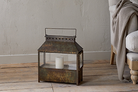 The Akoko Wide Lantern by nkuku, featuring a brown patina and glass panels, holds a white candle on a wooden floor next to a beige upholstered chair with a gray knitted blanket against a textured light gray wall.