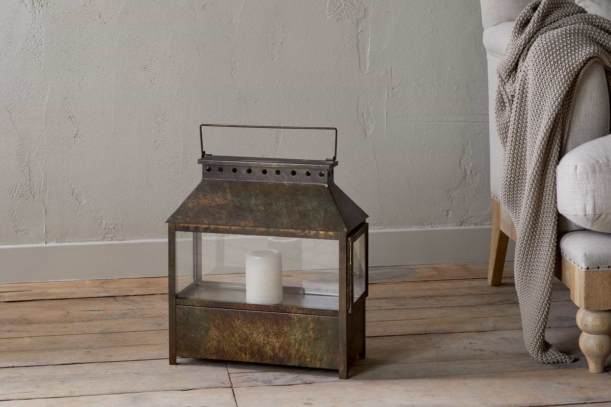 The Akoko Wide Lantern by nkuku, featuring a brown patina and glass panels, holds a white candle on a wooden floor next to a beige upholstered chair with a gray knitted blanket against a textured light gray wall.