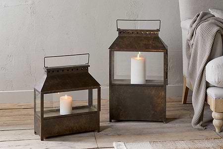 Two square Akoko Wide Lanterns by nkuku, finished in brown patina and each holding a lit white pillar candle, rest on a wooden floor. A beige armchair with wooden legs and a knitted throw sits against a textured neutral wall.
