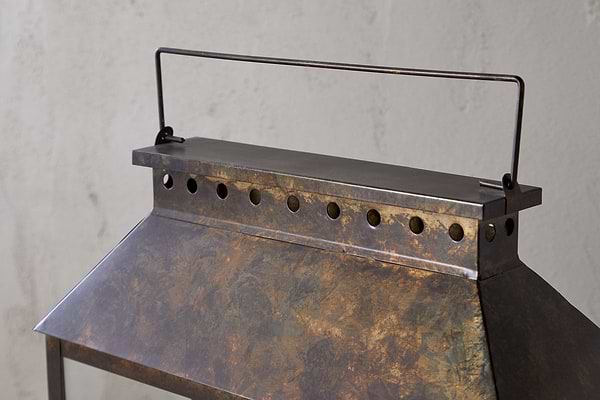 Close-up of the Akoko Wide Lantern by nkuku, featuring an oxidized brown patina, circular ventilation holes, and a slim black handle. The lantern stands out against a plain, lightly textured white wall.