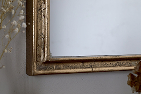 Close-up of the Almora Wall Mirror - Antique Brass (Extra Small) by nkuku, showing its slightly distressed frame against a light wall. Delicate dried flowers appear at the left and bottom edges.