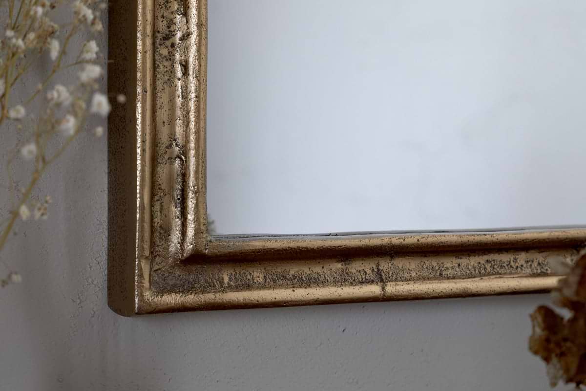 Close-up of the Almora Wall Mirror - Antique Brass (Extra Small) by nkuku, showing its slightly distressed frame against a light wall. Delicate dried flowers appear at the left and bottom edges.