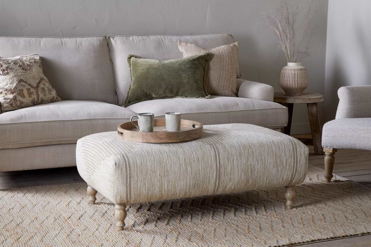 A cozy living room featuring a beige sofa, two accent pillows, the Aranya Upholstered Ottoman - Natural by nkuku, a round wooden tray with two mugs, textured rug, side table, and a ceramic vase with dried grasses for the perfect touch.