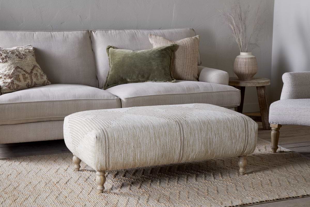 A cozy living room features a beige sofa with textured pillows, a matching armchair, and the nkuku Aranya Upholstered Ottoman - Natural as the perfect accent. A woven rug adds warmth, while a wooden side table displays a ceramic vase with dried grasses.