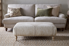 A neutral-toned living room features a light beige sofa with three textured pillows, paired with the Aranya Upholstered Ottoman – Natural by nkuku on a woven rug—a perfect living room accent.