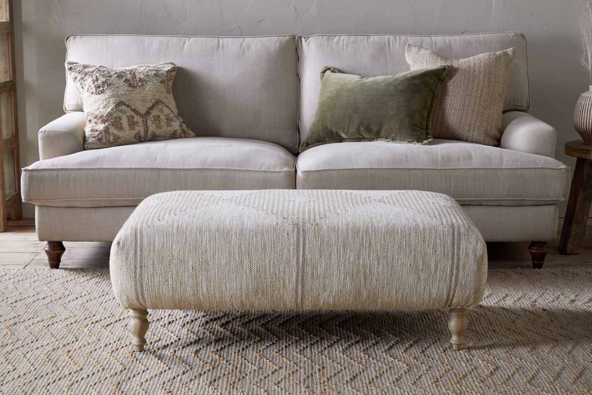 A neutral-toned living room features a light beige sofa with three textured pillows, paired with the Aranya Upholstered Ottoman – Natural by nkuku on a woven rug—a perfect living room accent.
