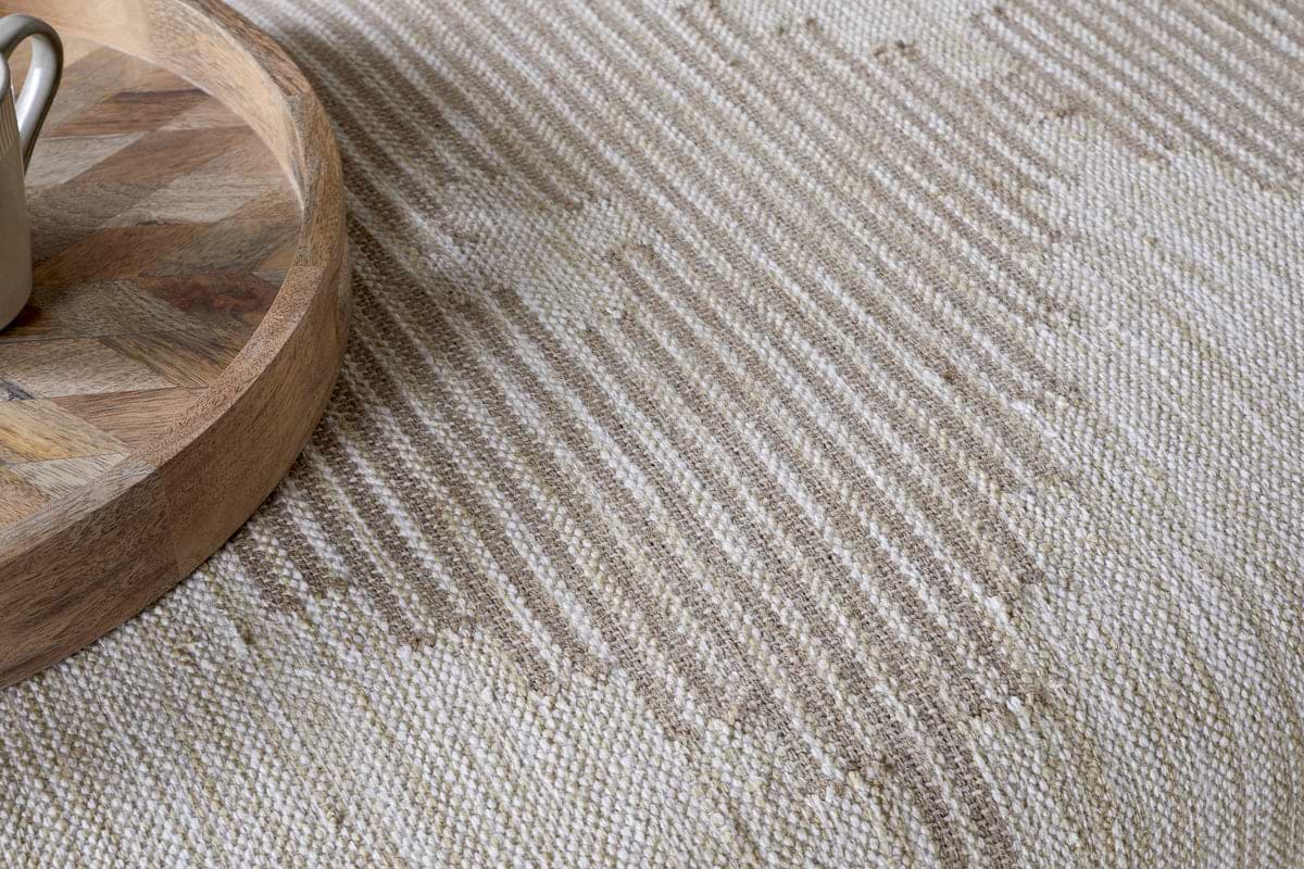 A close-up of the Aranya Upholstered Ottoman in Natural by nkuku showcases its textured beige and off-white zigzag fabric, ideal as a living room accent. A geometric wooden tray with a metal object appears partially in the corner.