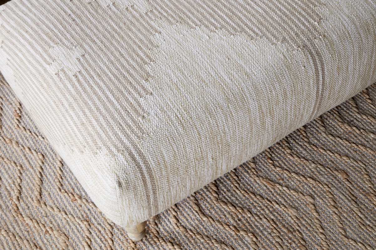 A close-up of the nkuku Aranya Upholstered Ottoman in Natural, featuring a subtle geometric pattern, sits on a tan and brown zigzag rug—making it an ideal stylish accent for your living room.