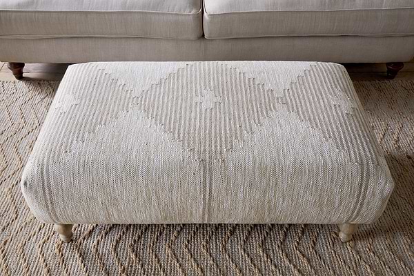 The Aranya Upholstered Ottoman - Natural by nkuku adds style to your living room with its beige textured fabric, subtle diamond pattern, and short wooden legs, standing out as a chic accent in front of a light-colored sofa.