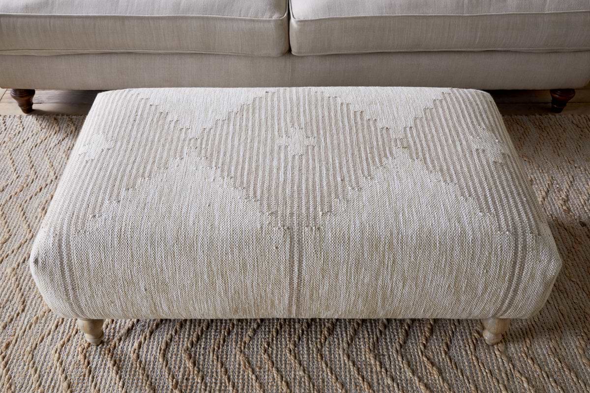 The Aranya Upholstered Ottoman - Natural by nkuku adds style to your living room with its beige textured fabric, subtle diamond pattern, and short wooden legs, standing out as a chic accent in front of a light-colored sofa.
